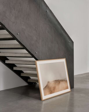 Framed art print depicting a mountain top in beige hues, in a oak frame, leaning against a concrete starcase