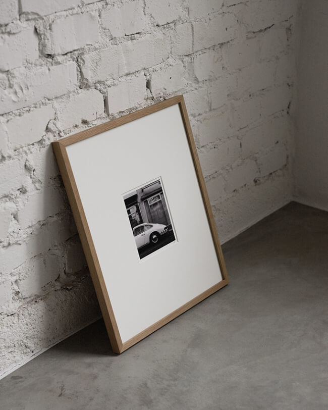 A framed art print leans against a white brick wall on a concrete floor. The artwork depicts a parked 911 porsche in black and white